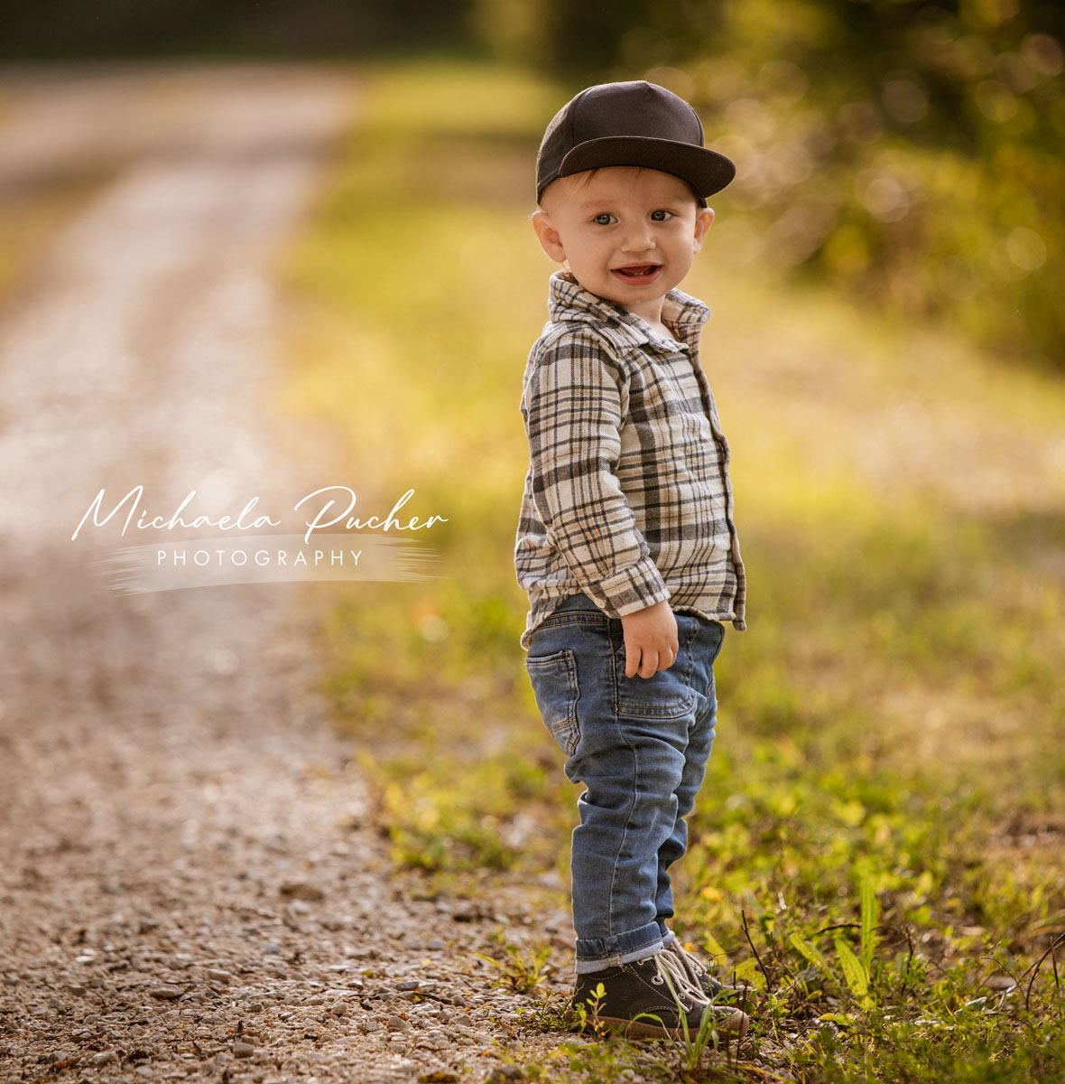 Kleiner Junge mit Kappe steht auf einem sonnigen Weg, lächelt neugierig in die Kamera und trägt ein kariertes Hemd und Jeans.